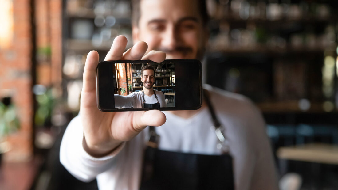 Smiling restaurant owner holding smartphone with camera on make selfie